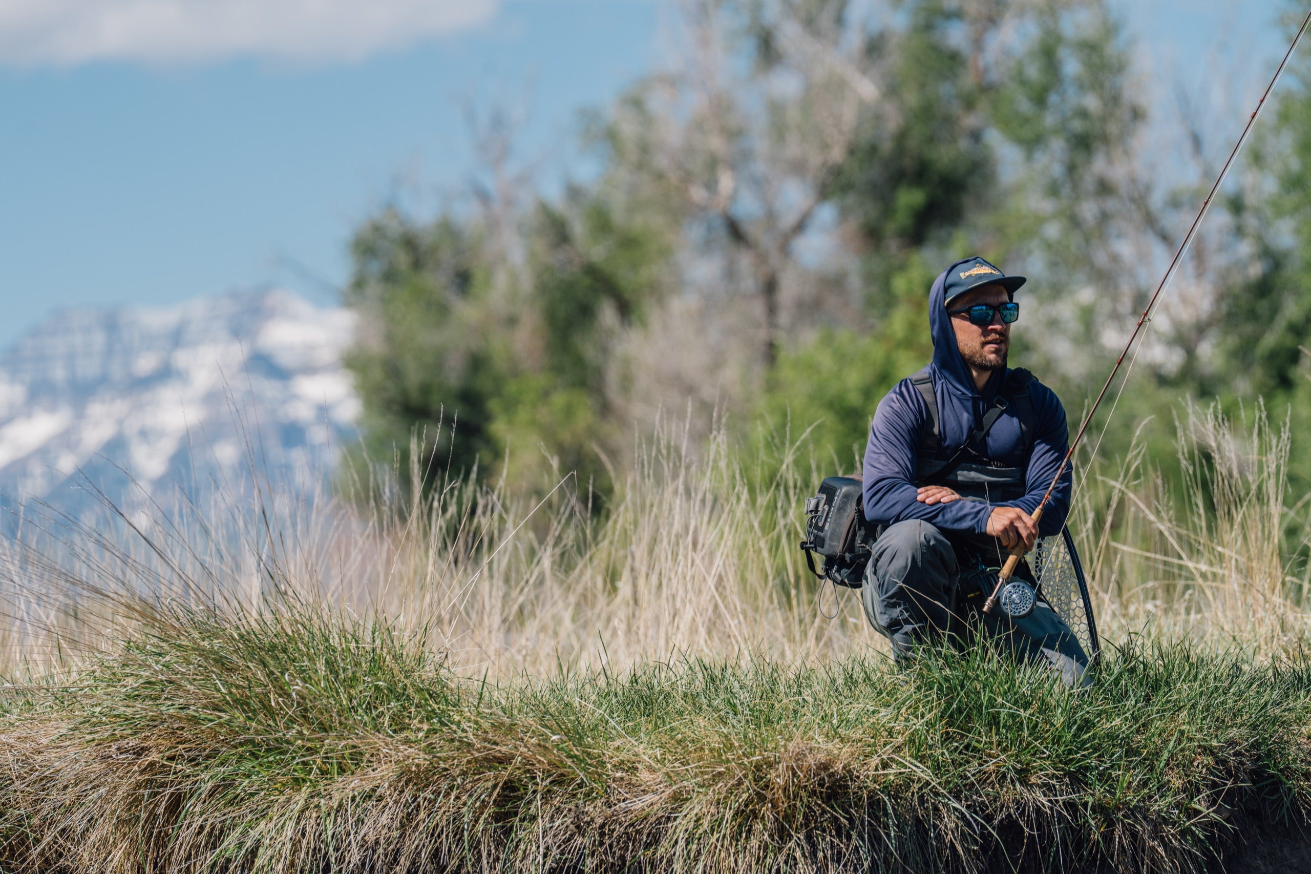 Angler kneeling in tall riverside grass with mountains behind